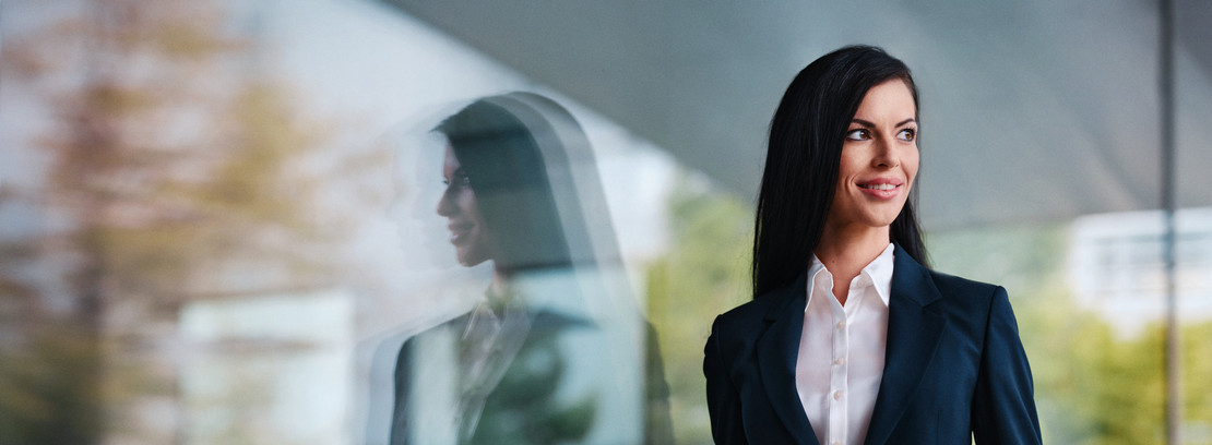 Une jeune courtière se tient devant un bâtiment administratif vitré et regarde par-dessus son épaule en souriant, tandis que son reflet apparaît dans la vitre