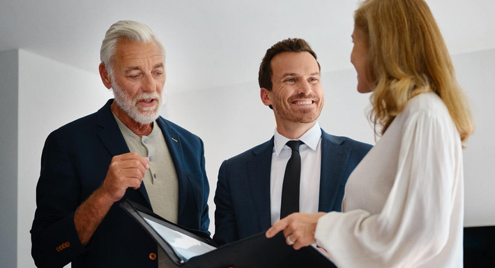 À l’intérieur, un courtier souriant se tient debout entre un couple d’un certain âge et lui présente des documents de vente