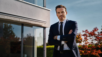 Selbstbewusster Makler sieht in die Kamera mit Haus im Hintergrund